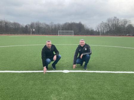 Dirk Tolkemitt und Feyhan Karadeniz hocken auf dem neuen Kunstrasenplatz am Schulzentrum Aspe.